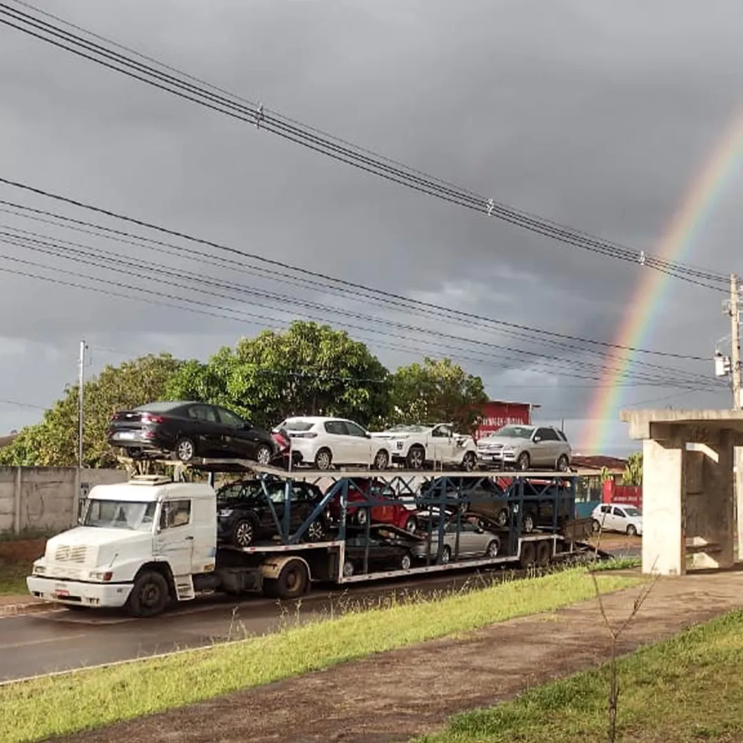 Cegonha da Capital carregada de veículos com arco-íris ao fundo